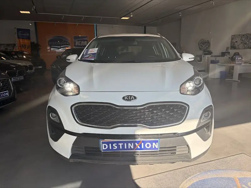 Vue frontale d’un Kia Sportage blanc 2021 en intérieur, avec calandre noire et phares allumés.