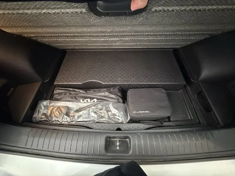 Vue du coffre ouvert d'un KIA Sportage blanc 2021, montrant le rangement sous plancher avec kit de secours et accessoires.