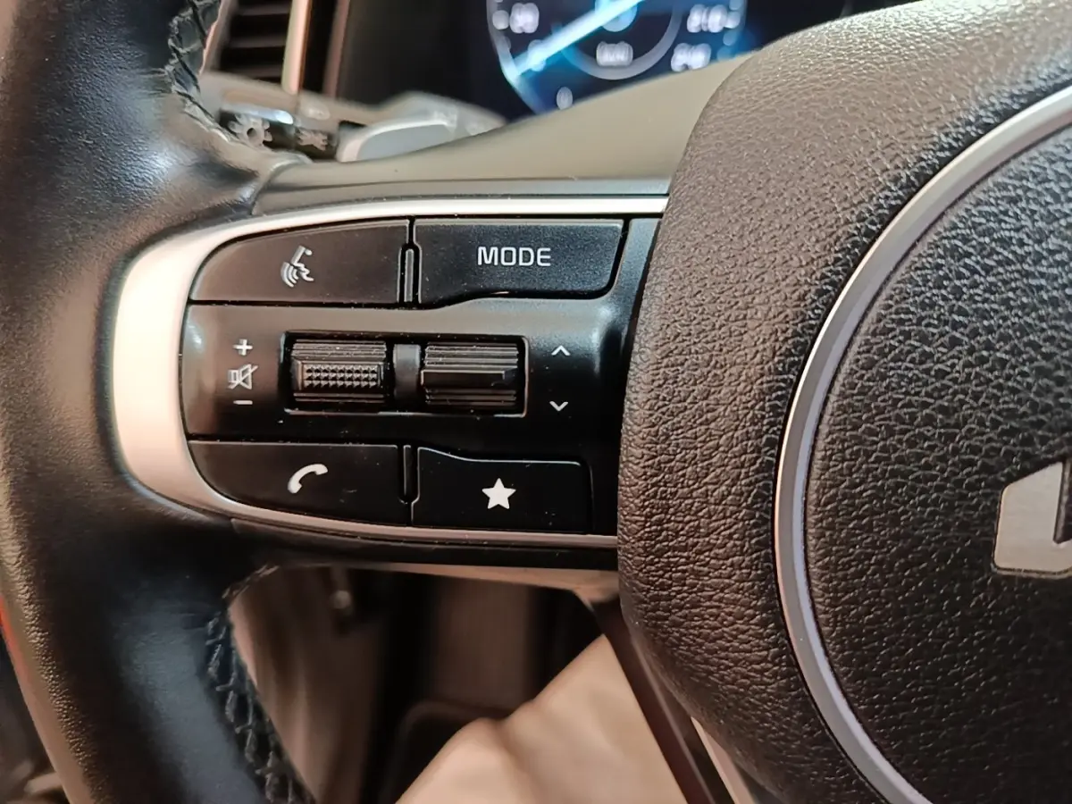 Gros plan sur les commandes gauche du volant cuir du Kia Sportage hybride rechargeable gris clair, avec affichage numérique en arrière-plan.