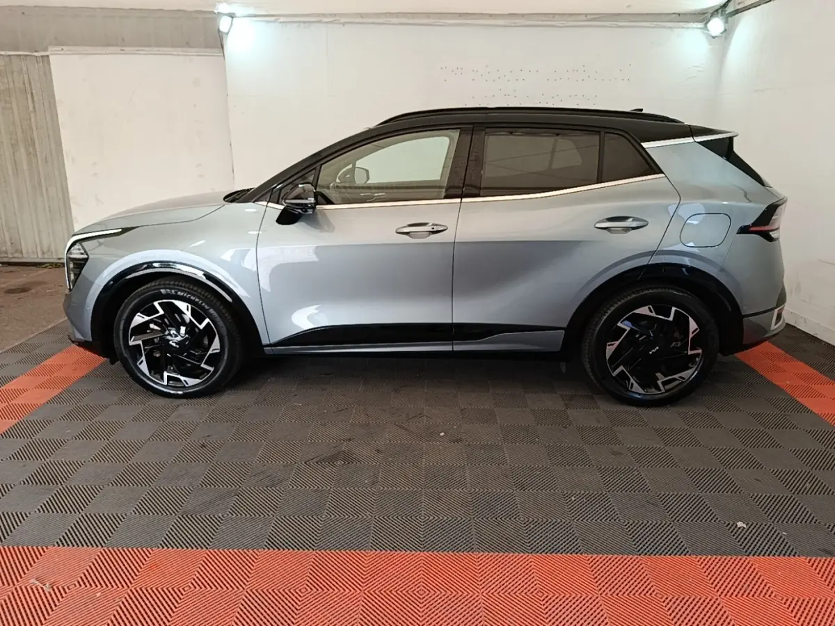 Vue de profil côté gauche d’un KIA Sportage gris clair avec toit noir et jantes noires à motifs argentés dans un garage.