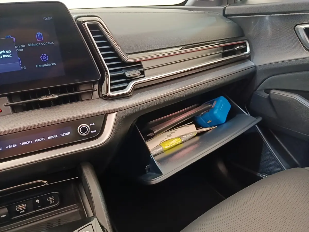 Intérieur côté passager du Kia Sportage 2022, tableau de bord noir avec boîte à gants ouverte contenant divers objets.