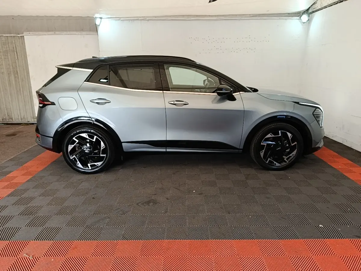 Vue de profil côté gauche d’un KIA Sportage gris clair avec toit noir et jantes noires stylisées dans un garage.