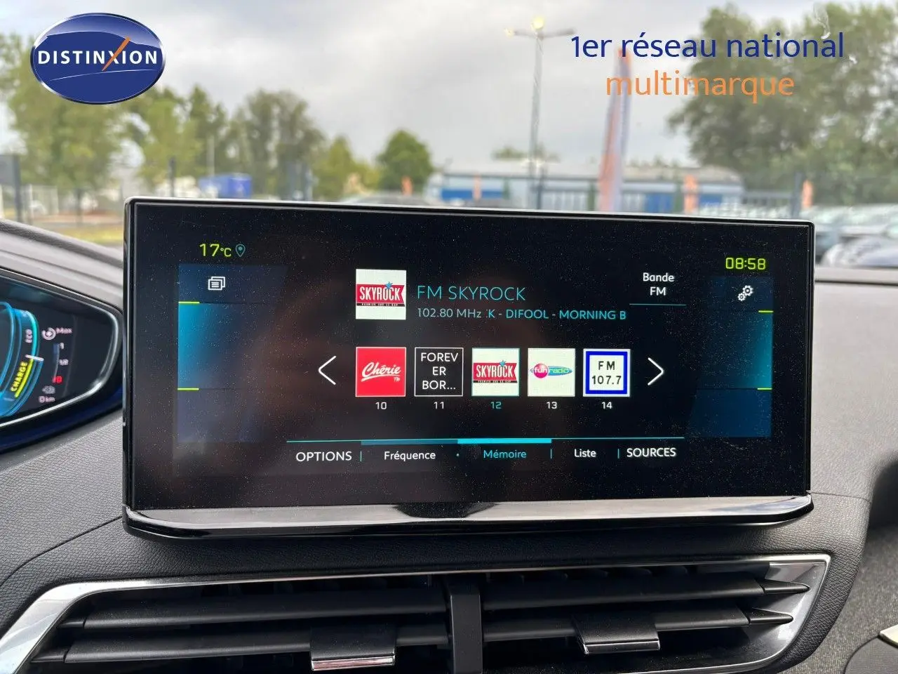 Écran tactile central du tableau de bord du Peugeot 3008 hybride, affichant les stations radio, avec vue sur l’extérieur flou.