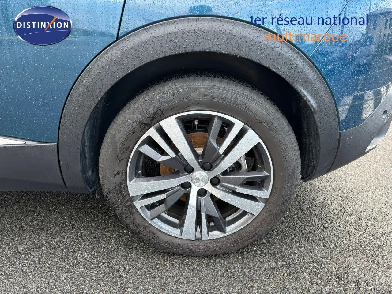 Gros plan sur la roue avant gauche et la jante biton noir et argent du Peugeot 3008 bleu Célèbes métal sous la pluie.