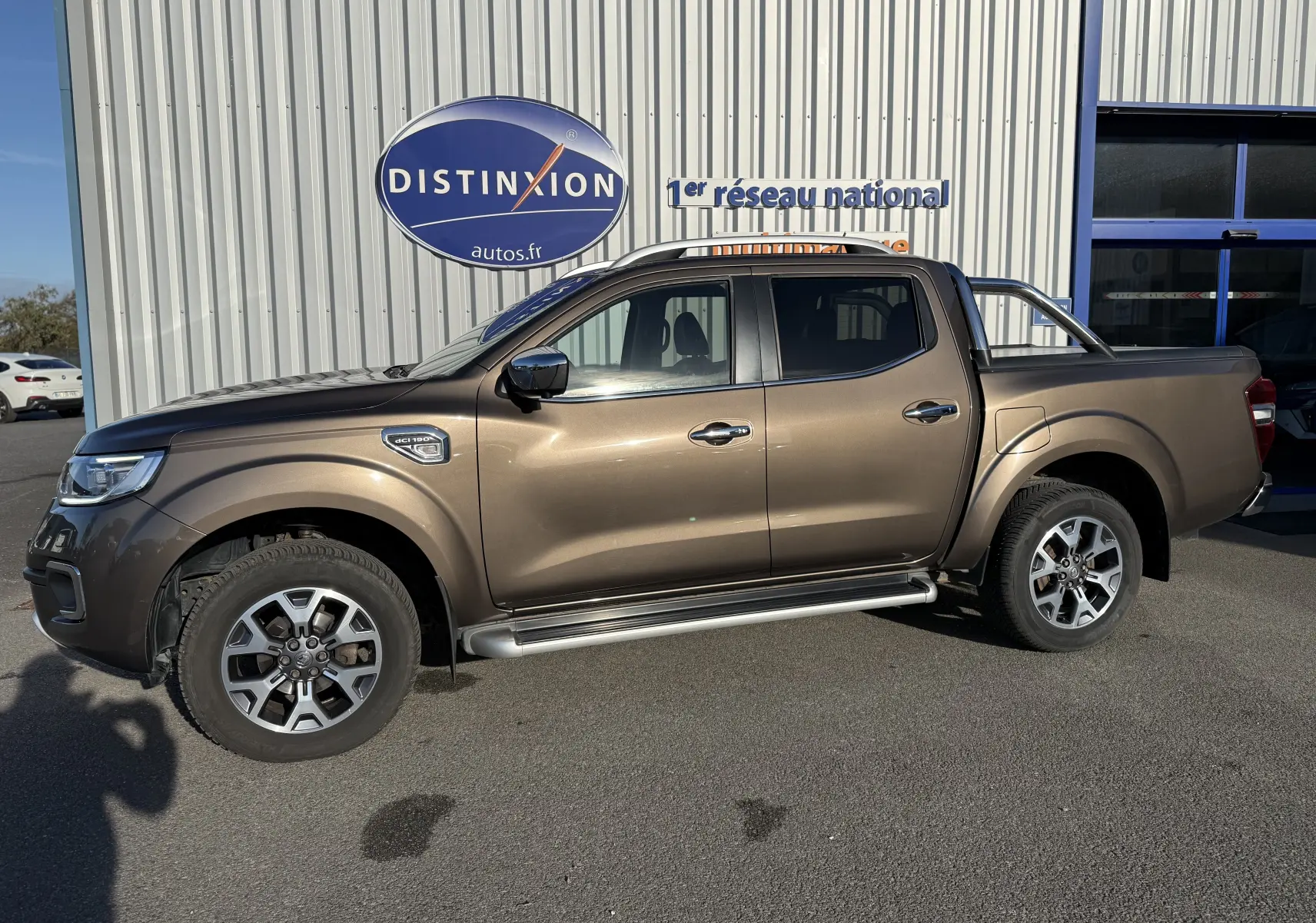 Renault Alaskan brun 2018 en 3/4 avant droit, avec calandre chromée et phares LED visibles, stationné devant un bâtiment.