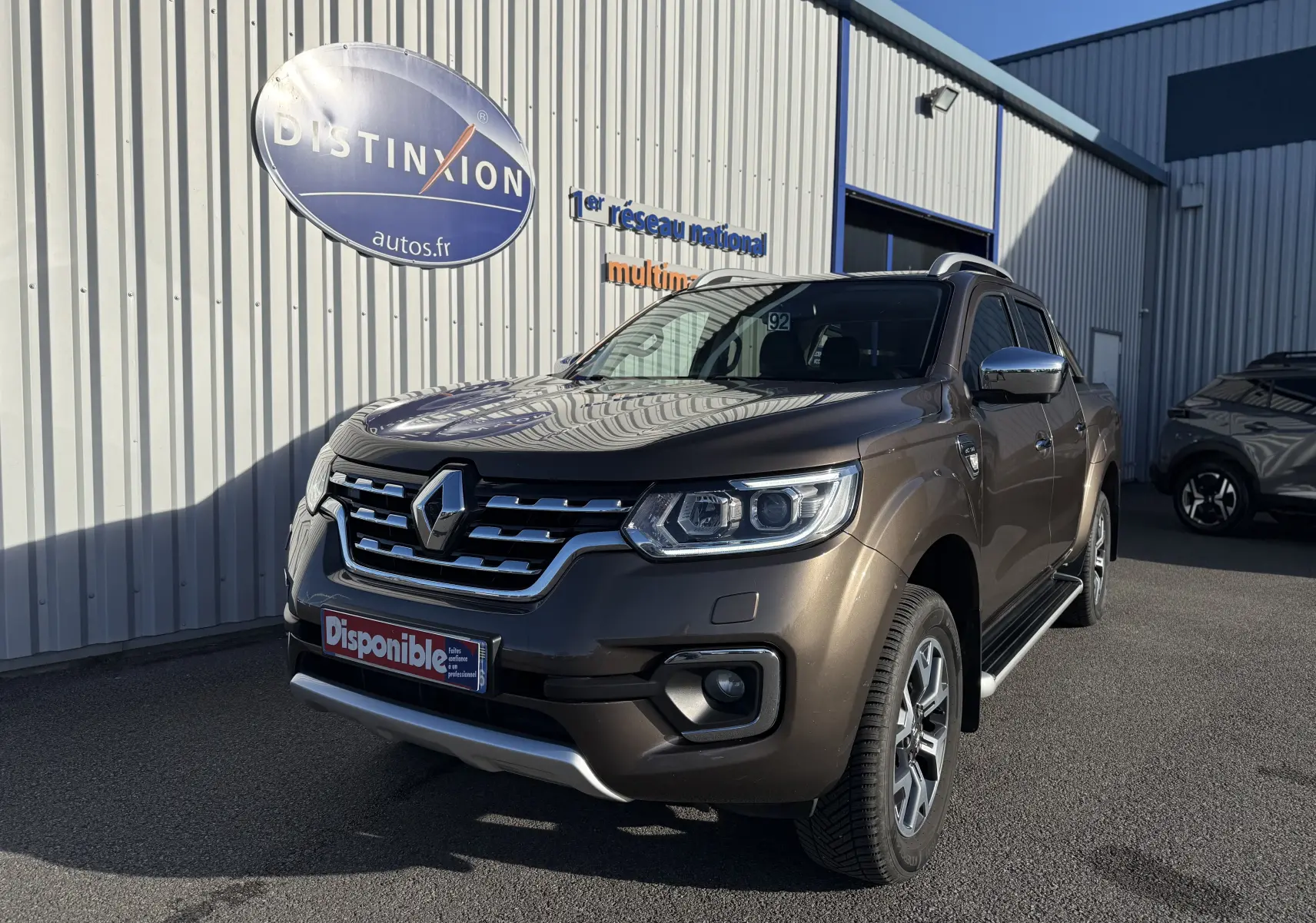 Profil côté gauche d'un pick-up Renault Alaskan brun 2018 avec double cabine et barres de toit, stationné devant un mur blanc.