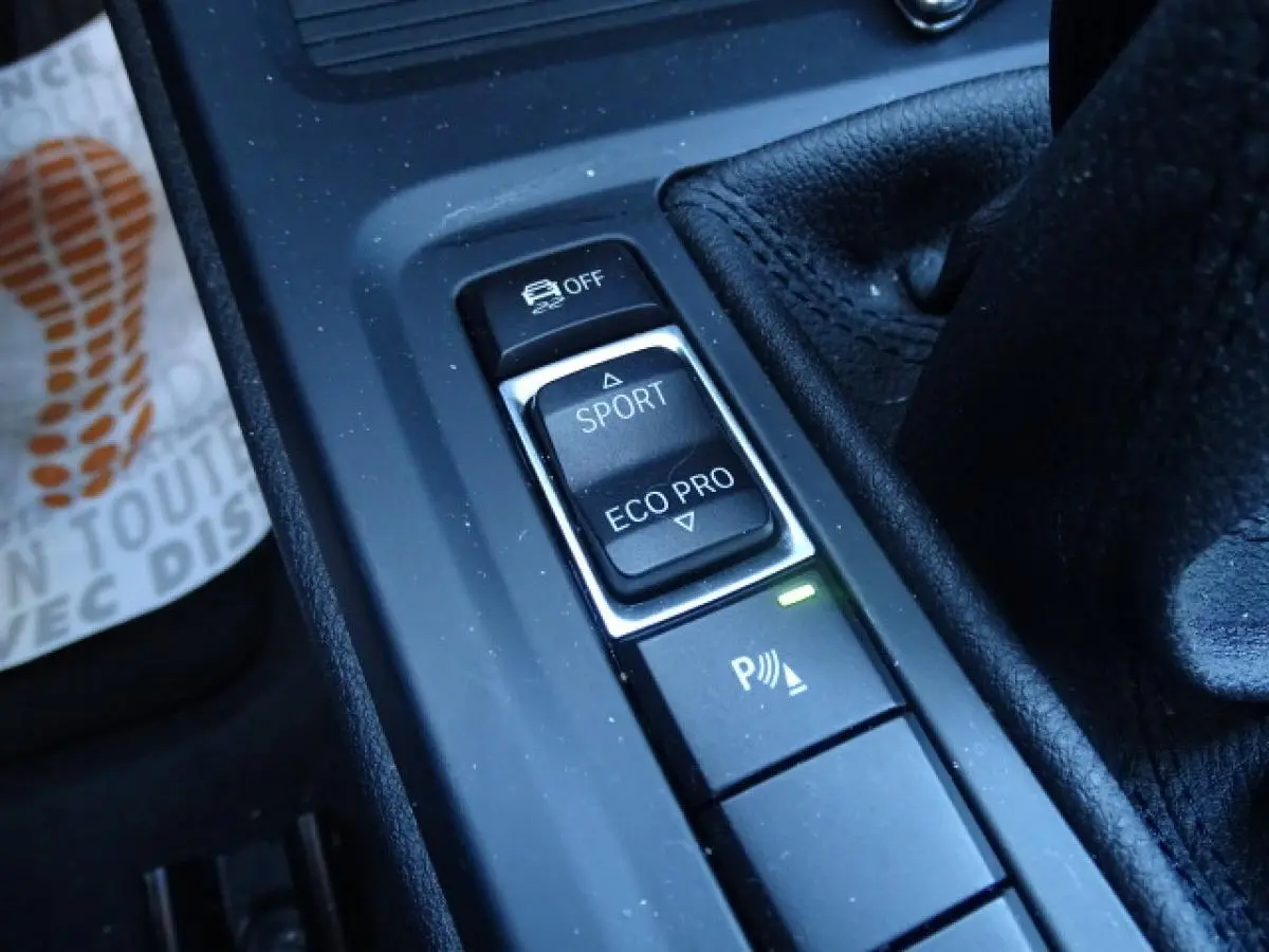 Gros plan sur les commandes de modes de conduite et assistance au freinage dans l’habitacle noir du BMW X1 blanc.
