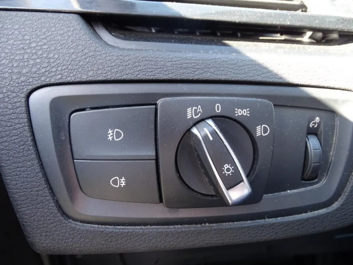 Gros plan sur le sélecteur de feux noir du tableau de bord du BMW X1 blanc, avec commandes d'éclairage et réglage de luminosité.
