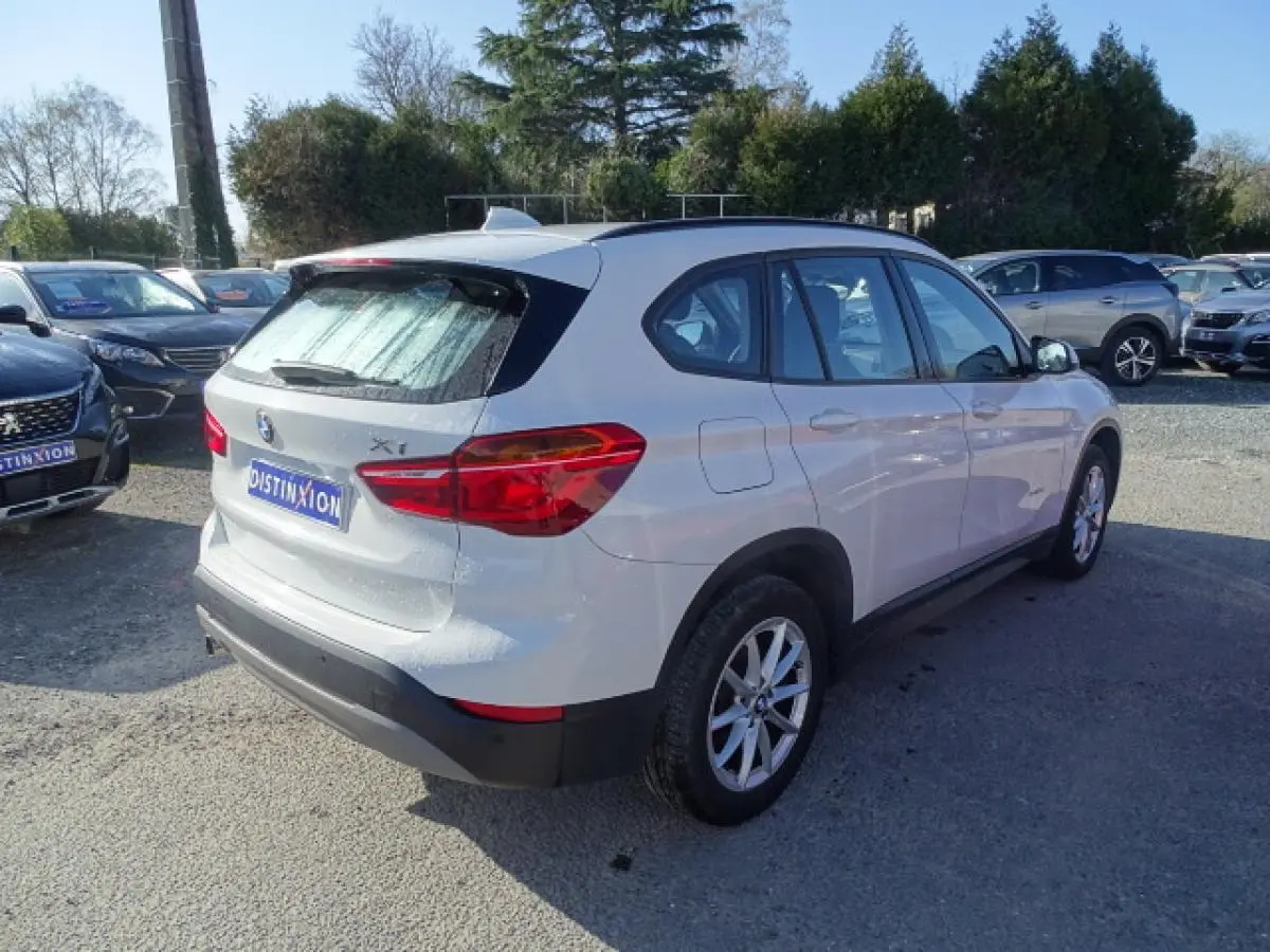 BMW X1 blanc vue 3/4 arrière droit sur parking, avec hayon et antenne aileron de requin visibles.