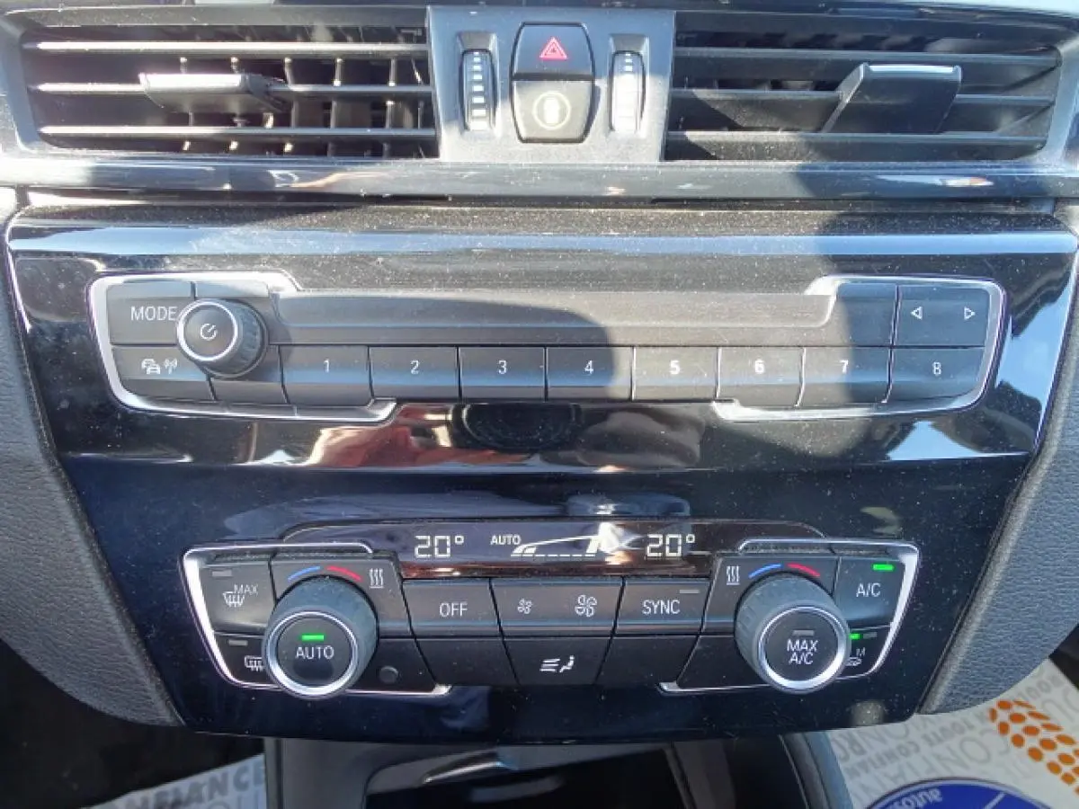 Vue rapprochée de la console centrale noire du BMW X1 blanc, montrant les commandes de climatisation automatique 2 zones.
