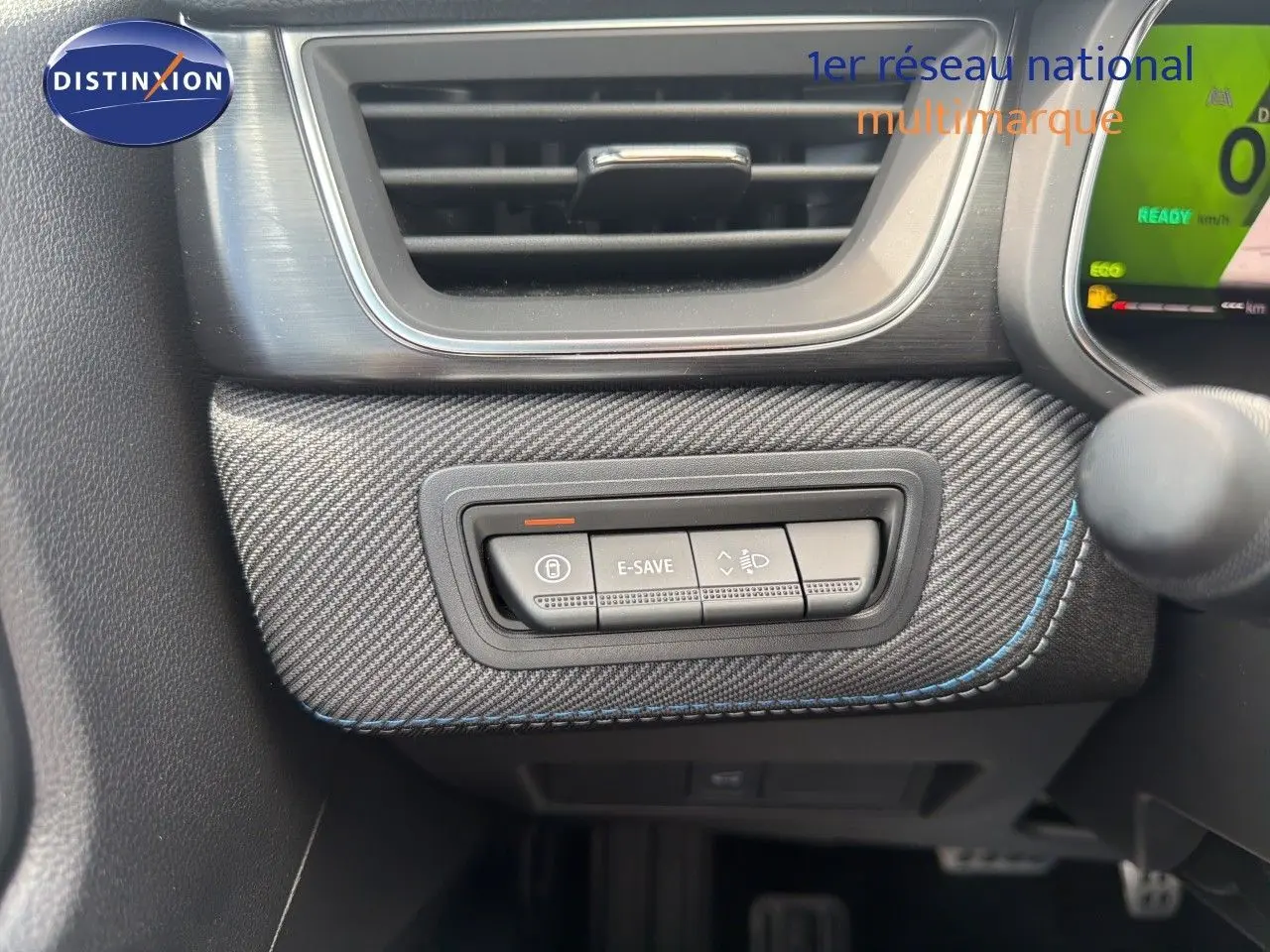Gros plan sur les commandes E-SAVE et réglage des phares, tableau de bord avec finition carbone et surpiqûres bleues du Renault Symbioz gris.