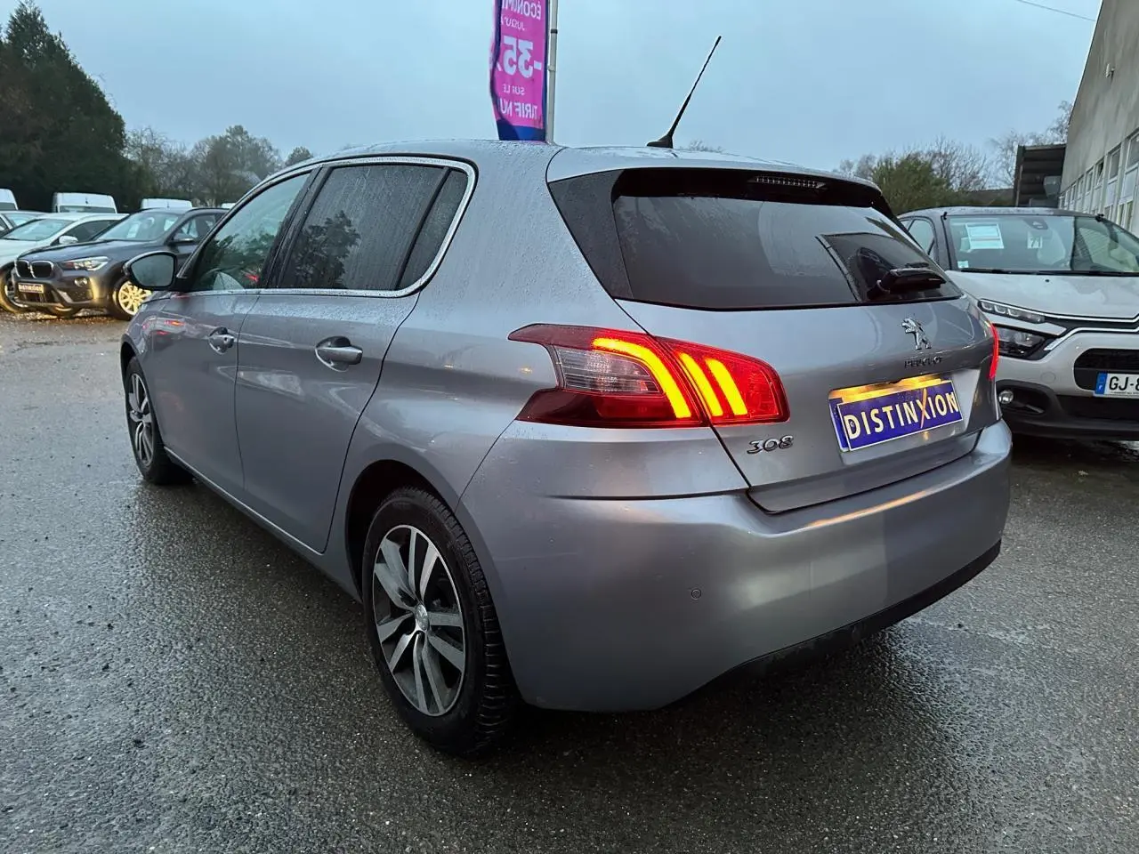 Vue 3/4 arrière gauche d'une Peugeot 308 gris Artense avec feux arrière allumés sur parking humide.