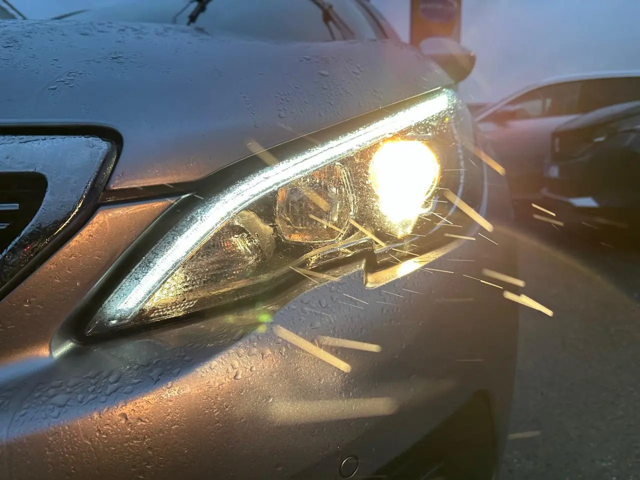 Gros plan sur le phare avant droit allumé d'une Peugeot 308 gris Artense avec gouttes d'eau au crépuscule.