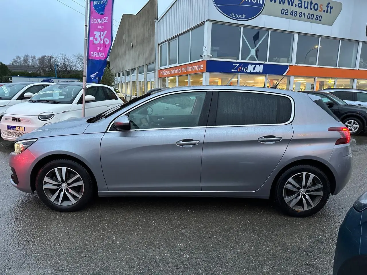 Profil côté gauche d'une Peugeot 308 gris Artense 2018 garée devant un concessionnaire sous la pluie.