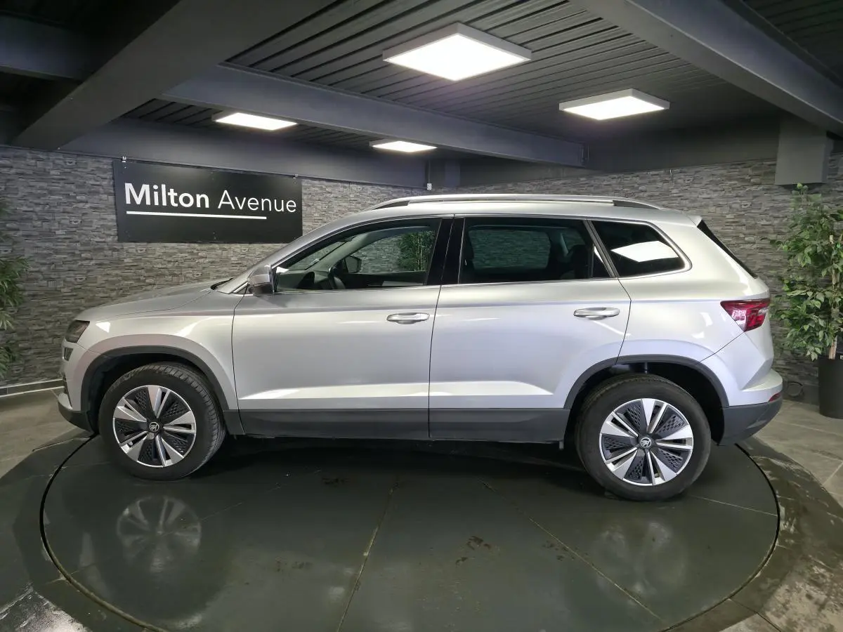 Vue latérale droite d'un SUV Skoda Karoq gris 2023 avec jantes alliage et barres de toit argentées dans un showroom.