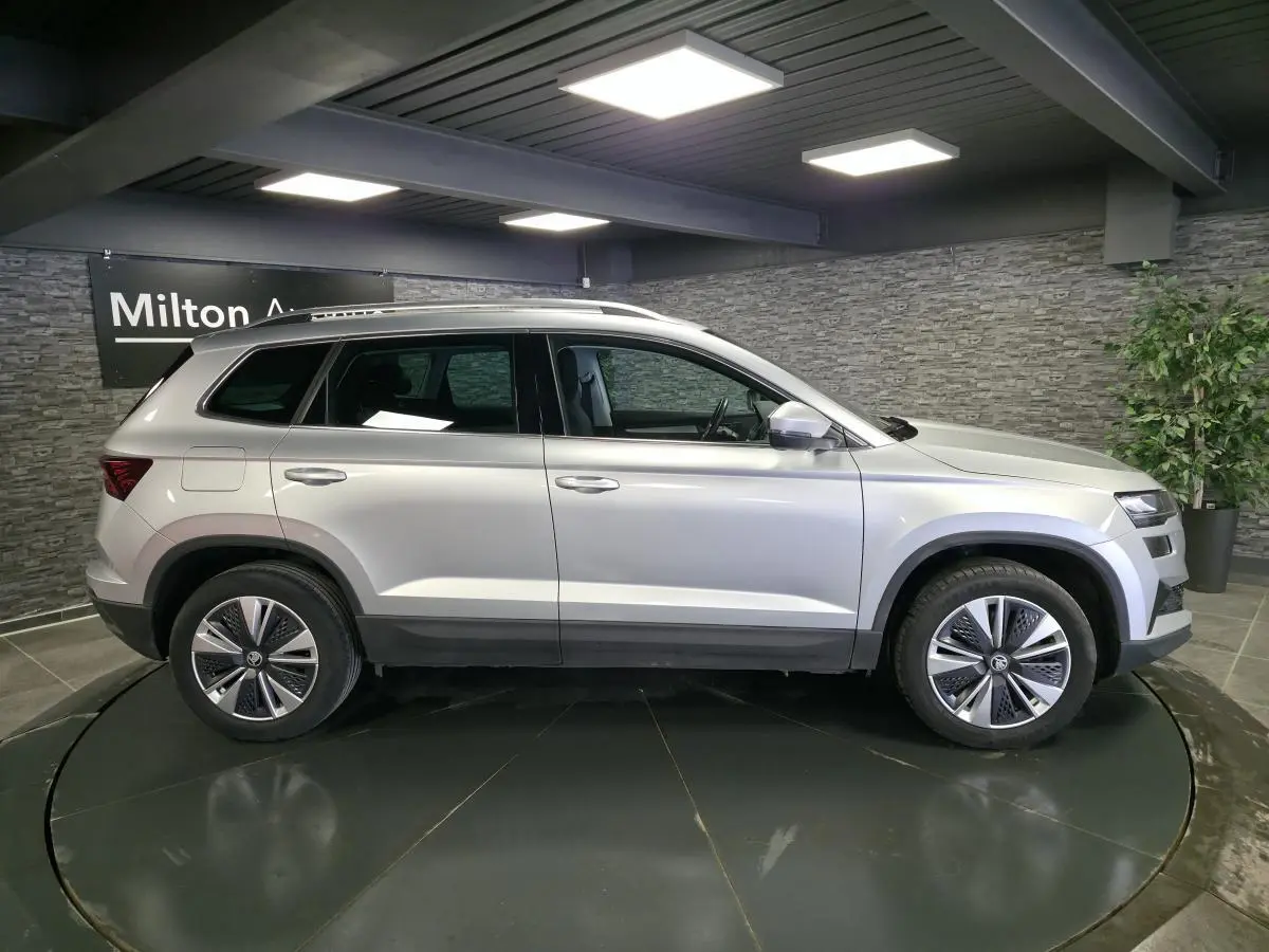 Vue latérale droite d'un SKODA KAROQ gris 2023 en intérieur avec jantes alliage et barres de toit noires.