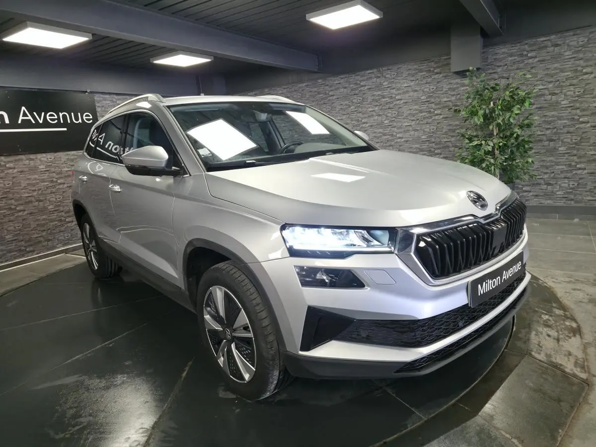 Skoda Karoq gris en 3/4 avant droit, phares allumés et calandre noire brillante dans un showroom.