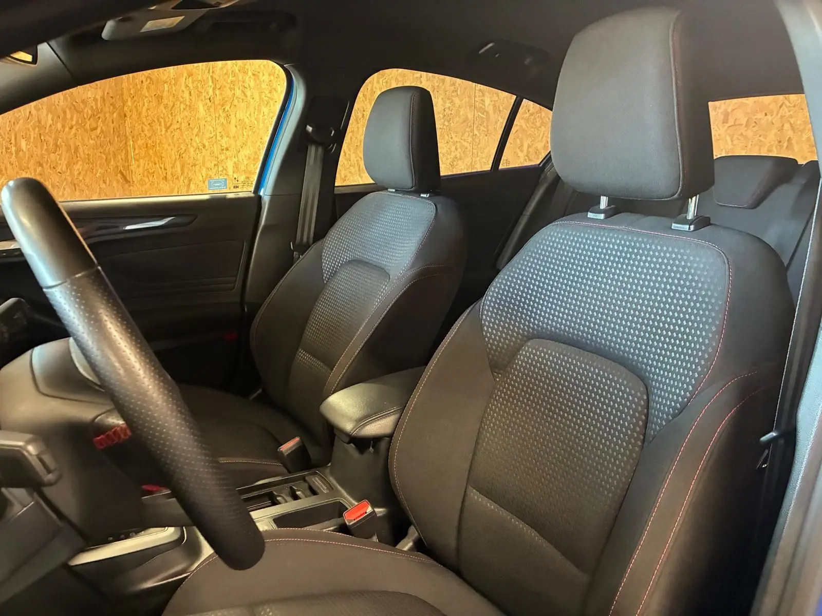 Vue intérieure côté avant gauche de la Ford Focus 2024, sièges tissu noir avec surpiqûres rouges et volant cuir perforé.