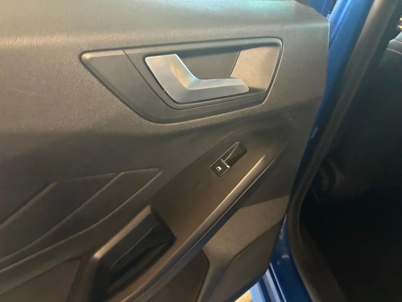 Détail de la porte intérieure côté gauche d'une Ford Focus bleu 2024, avec poignée argentée et bouton de vitre électrique.
