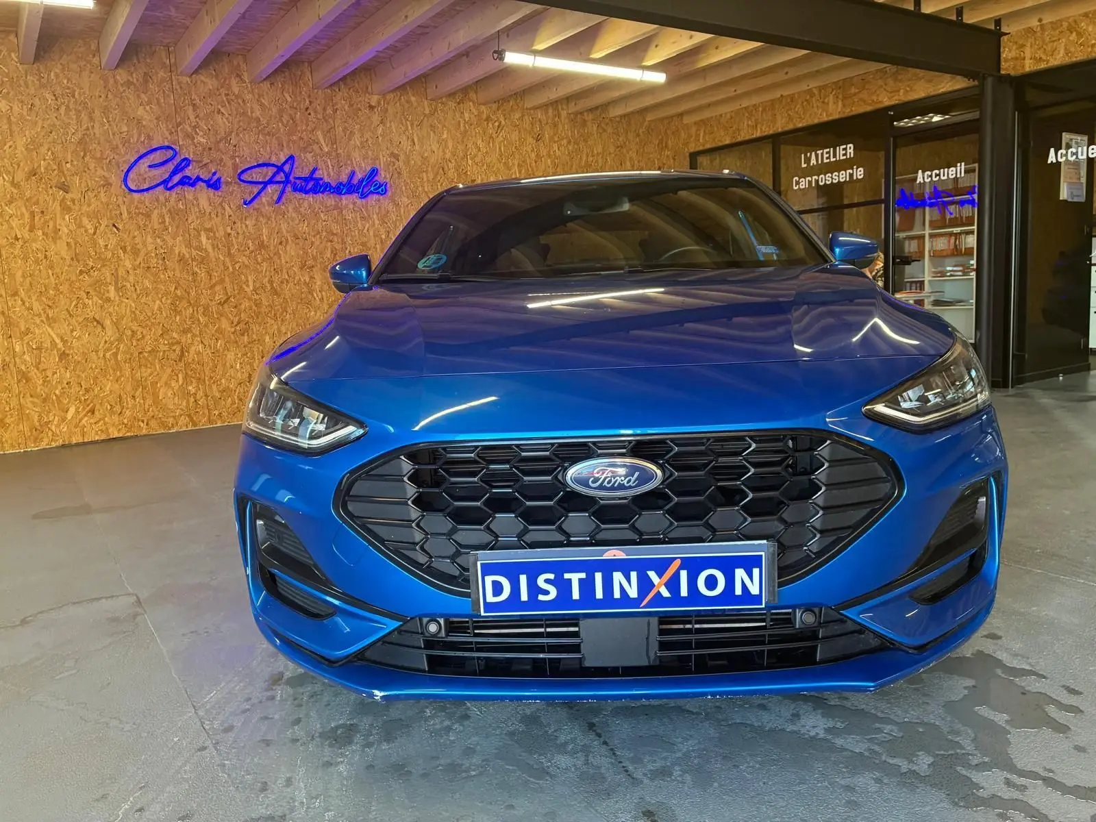 Vue frontale d'une Ford Focus bleue 2024 avec calandre noire hexagonale et plaque Distinxion en intérieur d'atelier.