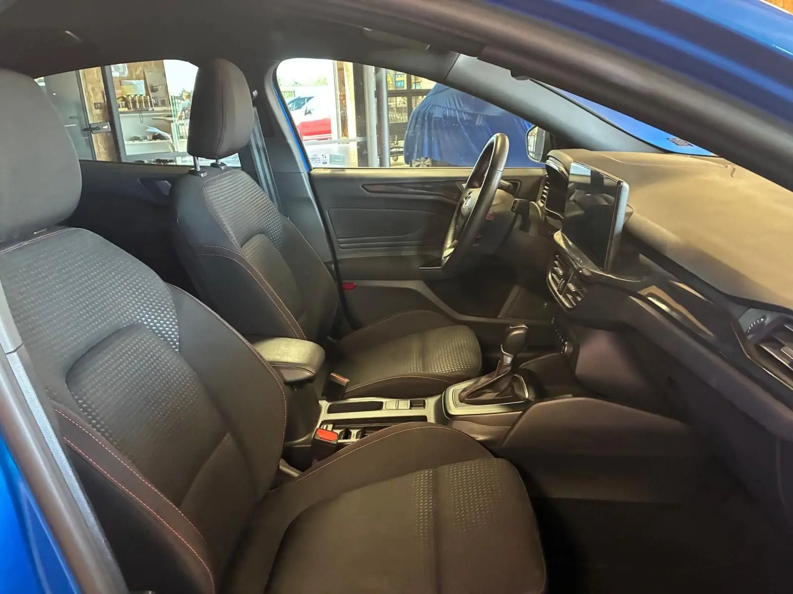 Vue intérieure côté gauche de la Ford Focus 2024 bleue, montrant sièges tissu noirs avec surpiqûres rouges et tableau de bord moderne.