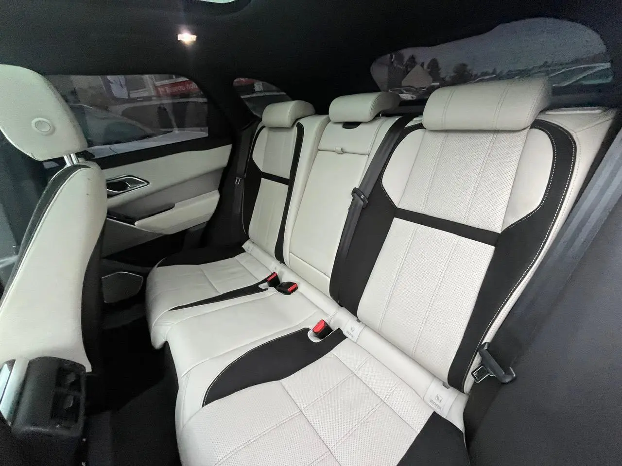 Vue intérieure côté droit sur la banquette arrière cuir blanc et noir du Range Rover Velar avec appuie-tête et ceintures visibles.