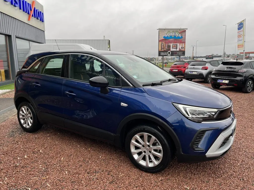 Opel Crossland X bleu clair en vue 3/4 avant droit, avec toit noir et jantes argentées, stationné sur un sol en gravier.