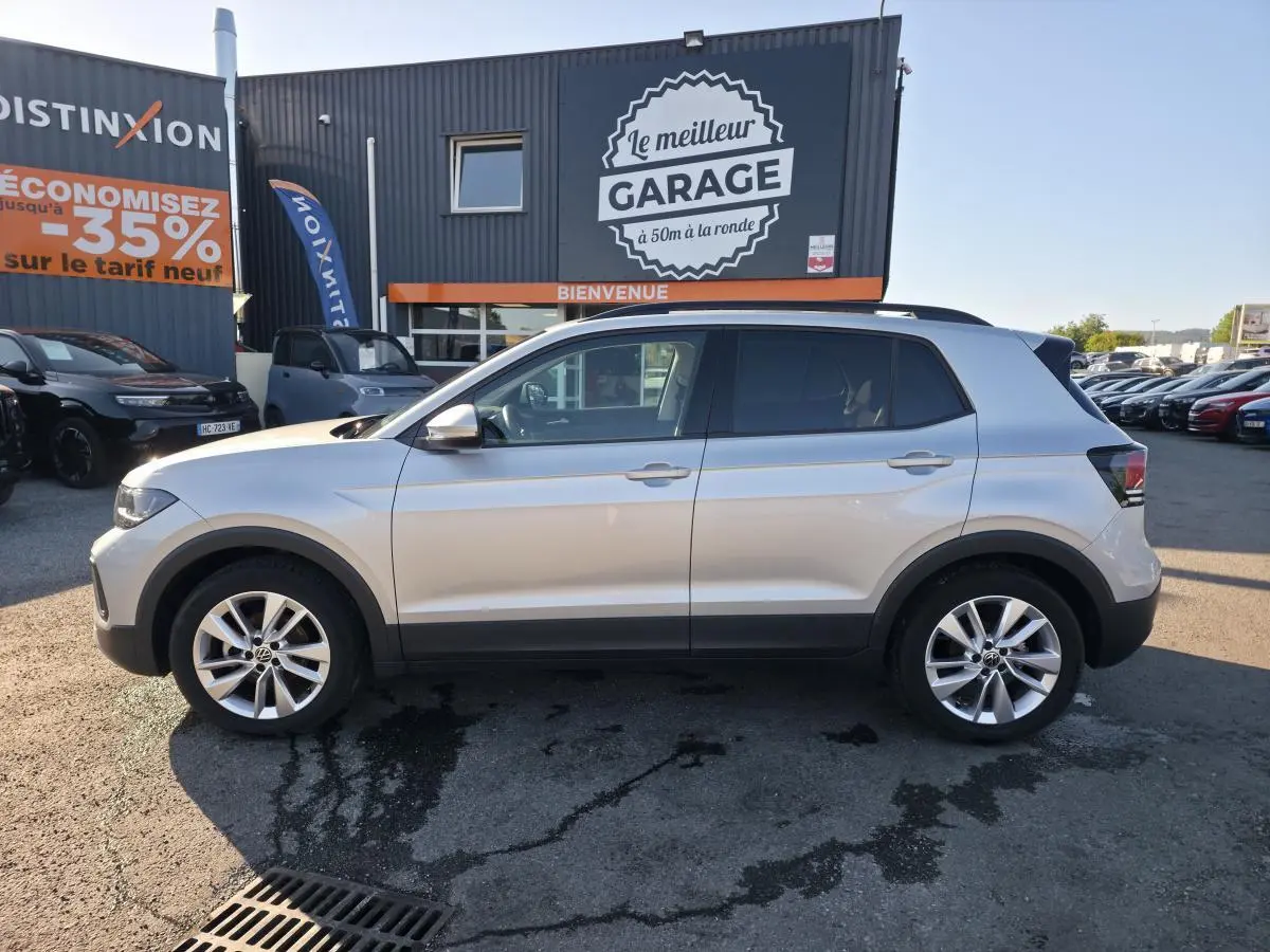 Profil côté gauche d'un Volkswagen T-Cross gris métallisé 2025 avec jantes alliage 17 pouces, stationné devant un garage.