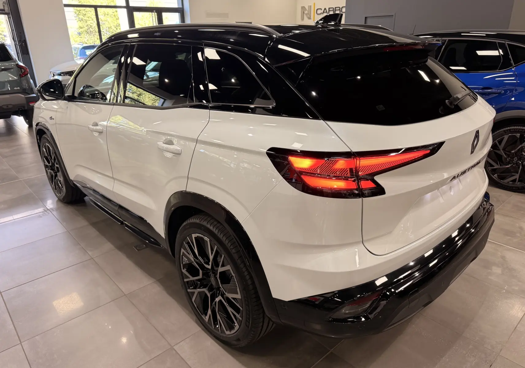 Vue 3/4 arrière droite du Renault Austral blanc nacré avec toit noir et feux arrière allumés en intérieur showroom.