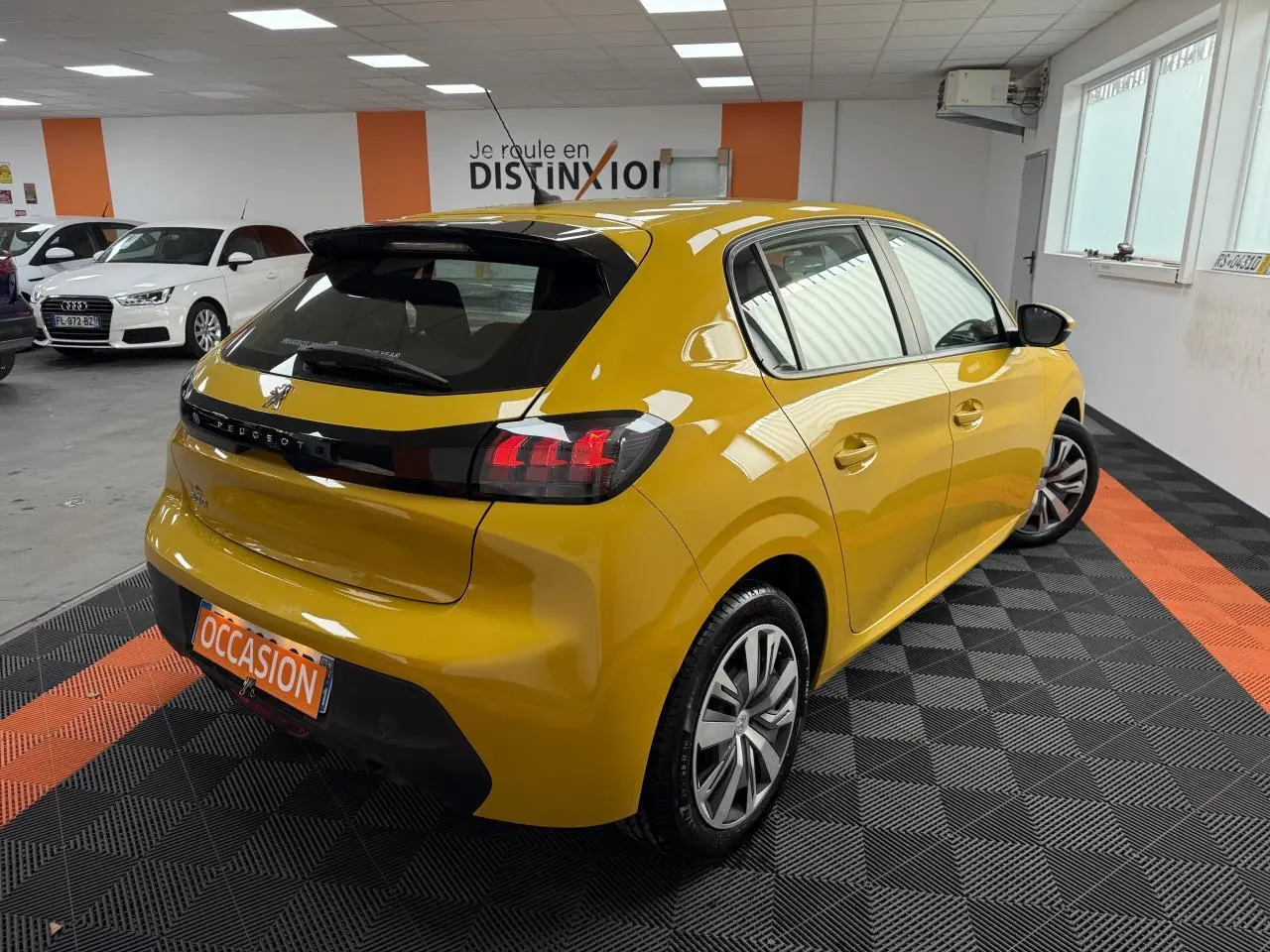 Peugeot 208 jaune clair vue 3/4 arrière droit en intérieur avec jantes 16 pouces et plaque occasion.