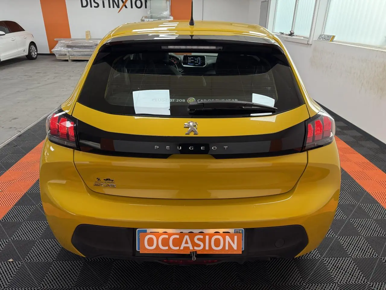 Vue arrière d'une Peugeot 208 jaune clair avec bande noire et plaque d'immatriculation orange "OCCASION" en intérieur showroom.
