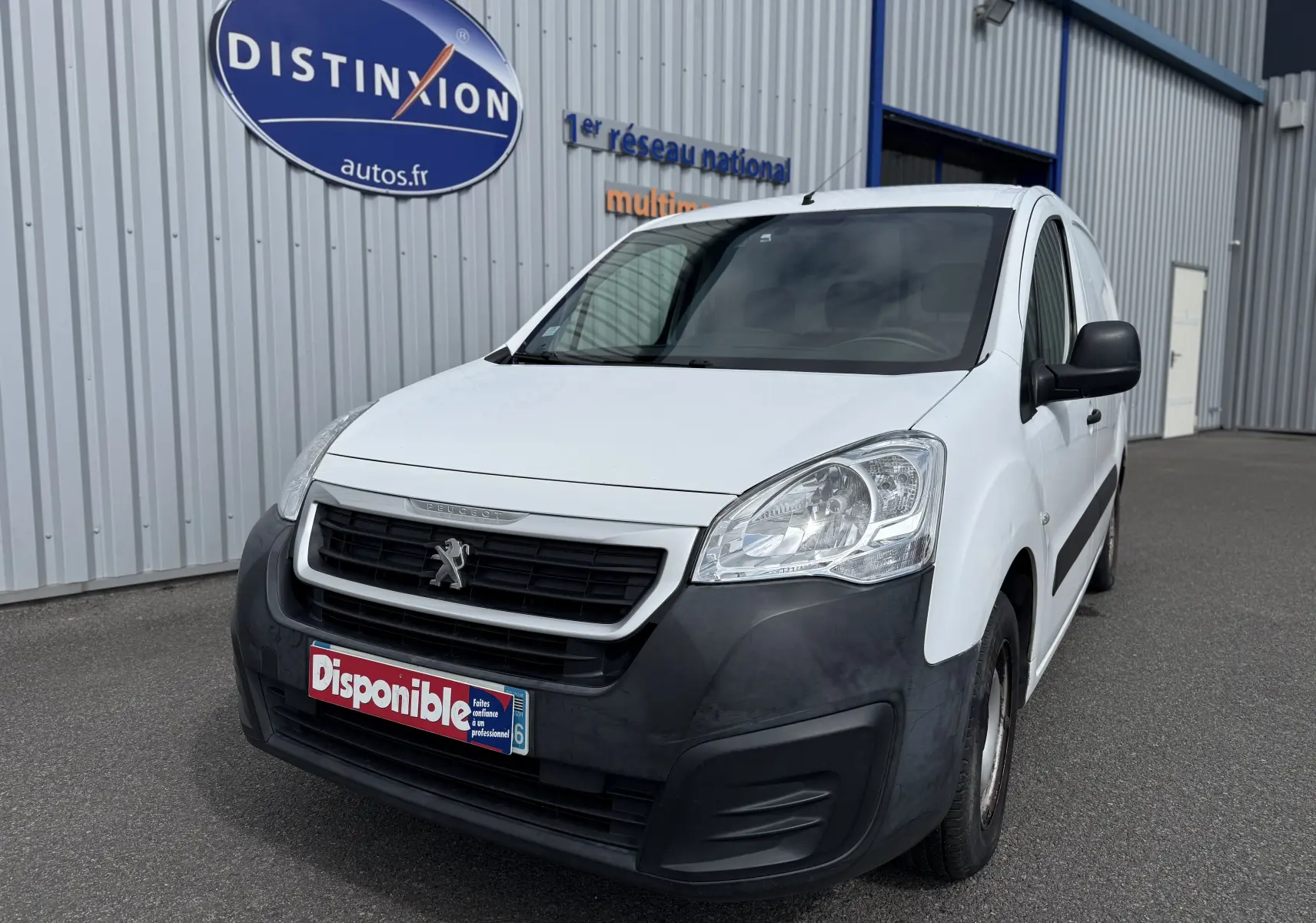 Peugeot Partner blanc vue 3/4 avant droit, avec pare-chocs noir mat et calandre distinctive, stationné devant un bâtiment industriel.