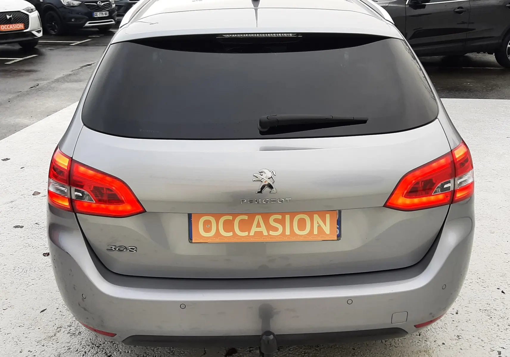 Vue arrière d'une Peugeot 308 SW gris Artense avec feux allumés et attelage visible sur un parking.