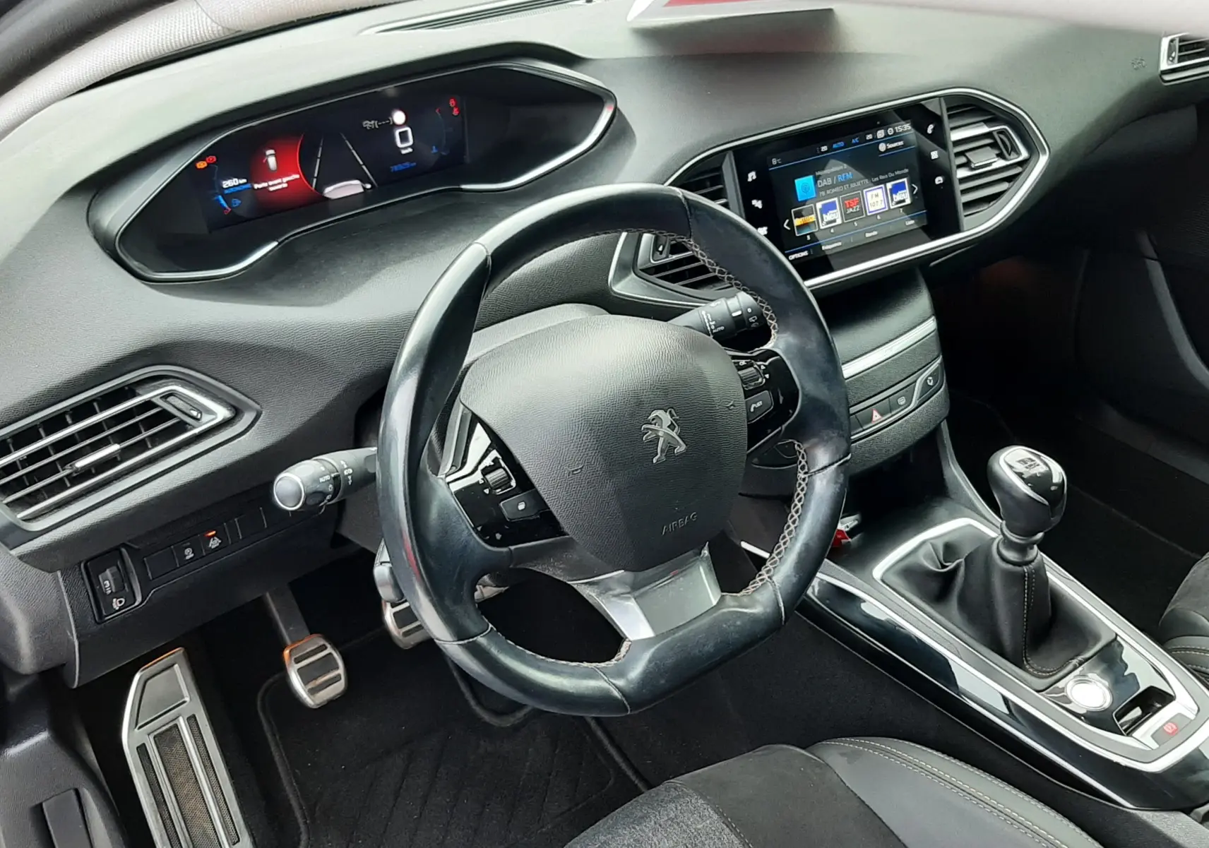 Vue intérieure centrée sur le volant Peugeot et le tableau de bord numérique avec écran tactile, sellerie tissu noir visible.