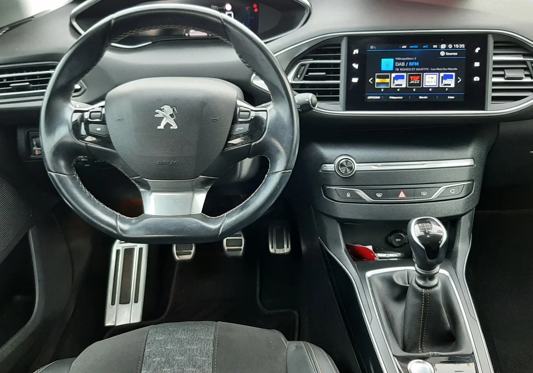 Vue intérieure frontale du poste de conduite de la Peugeot 308 SW 2021, volant cuir noir et écran tactile central allumé.