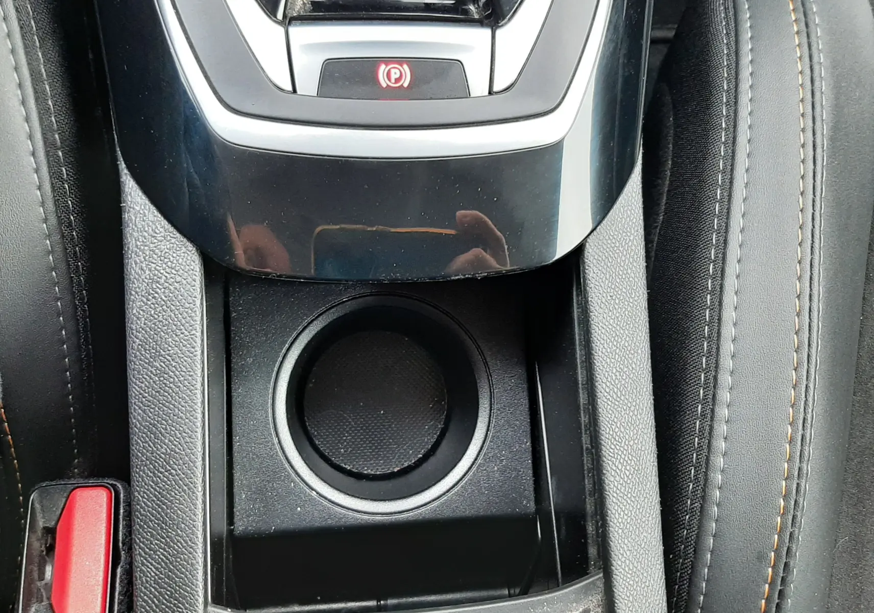 Vue rapprochée de la console centrale noire avec bouton de frein à main électrique et porte-gobelet, sièges tissu gris avec surpiqûres.