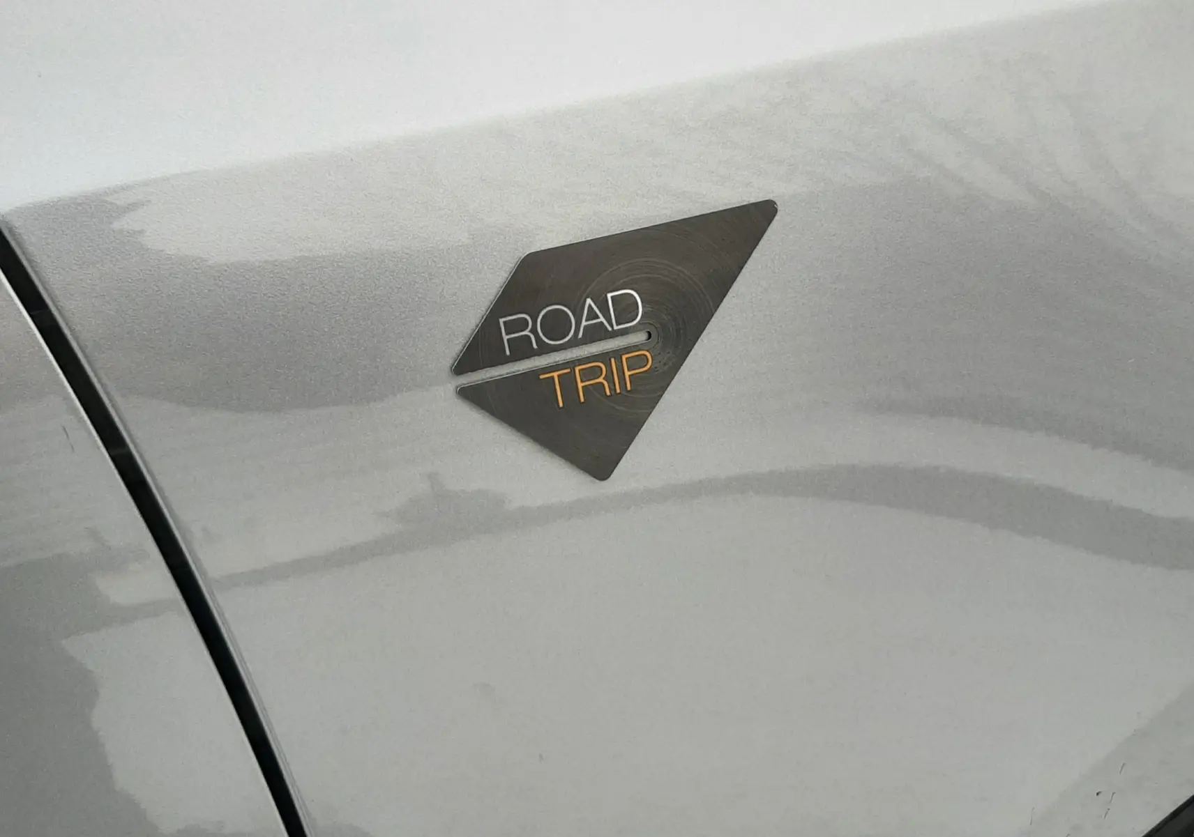 Gros plan sur l'écusson Road Trip noir et orange sur la carrosserie gris Artense d'une Peugeot 308 SW.