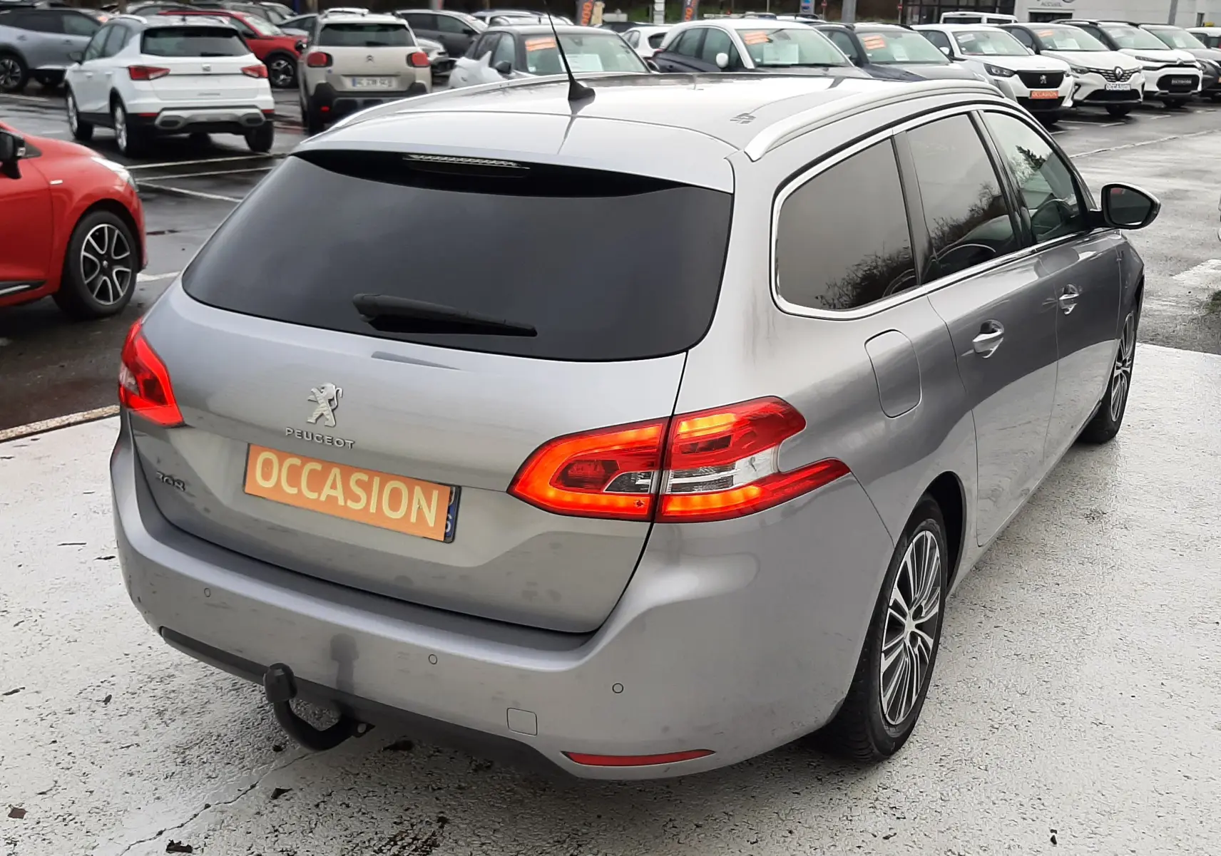 Vue 3/4 arrière droite d'une Peugeot 308 SW gris Artense avec feux allumés et attelage visible sur parking.