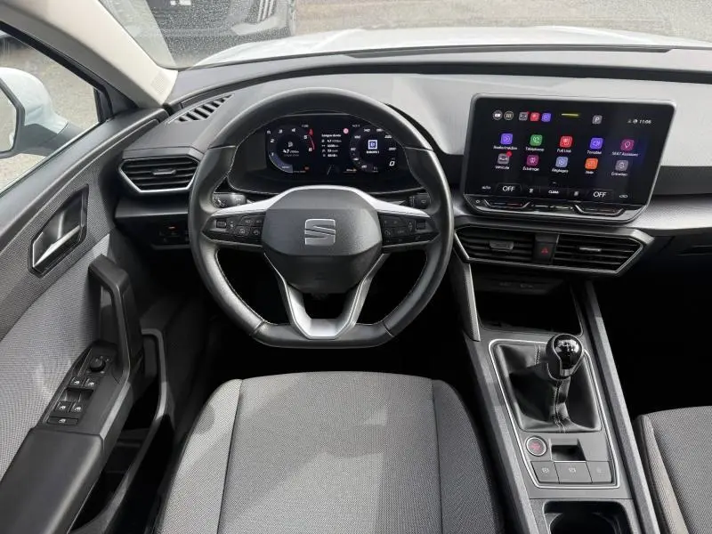 Vue intérieure frontale du poste de conduite du SEAT Leon 2025 blanc, volant cuir multifonction et écran tactile central.