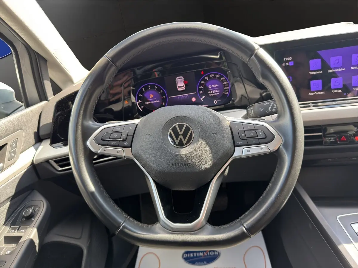Vue intérieure centrée sur le volant cuir noir et les écrans digitaux du tableau de bord de la Volkswagen Golf gris clair.