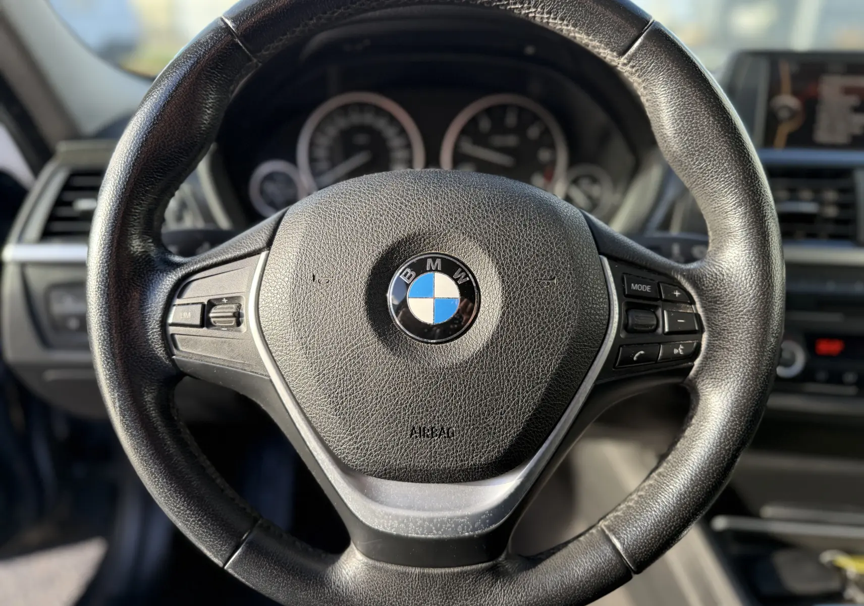 Gros plan sur le volant en cuir noir d'une BMW Série 3 320D gris, avec commandes intégrées et tableau de bord flou en arrière-plan.