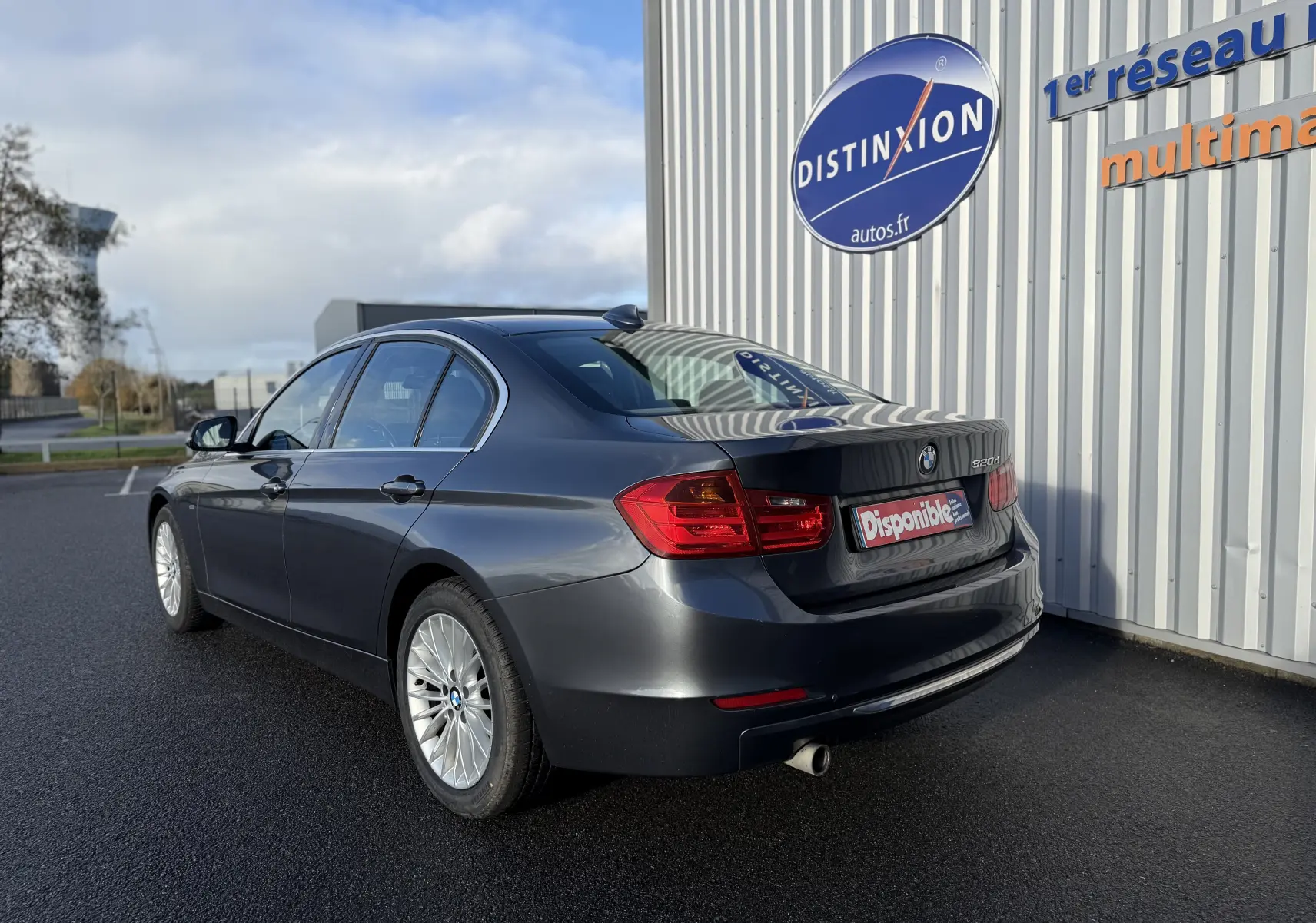 BMW Série 3 320D gris vue 3/4 arrière droit, avec jantes alliage et feu arrière rouge vif sous un ciel partiellement nuageux.