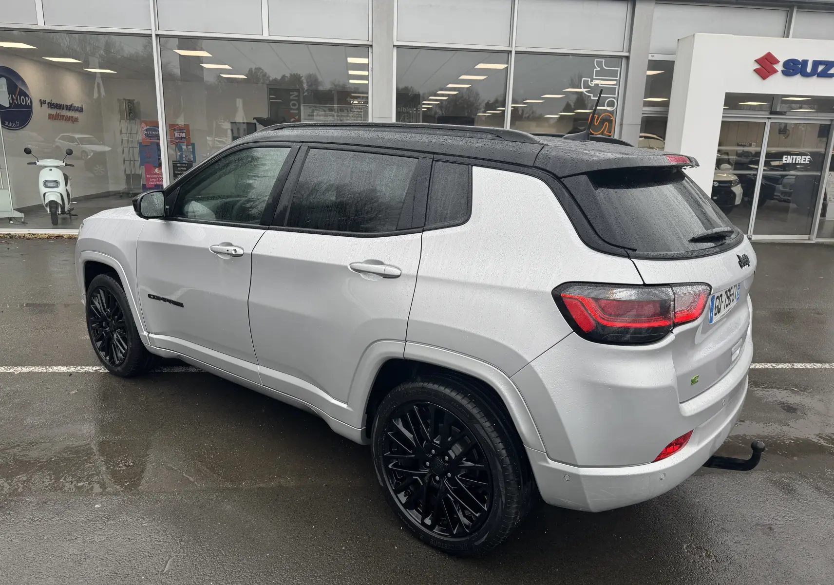 Jeep Compass gris clair vue 3/4 arrière droit avec toit noir et jantes noires brillantes sous la pluie.