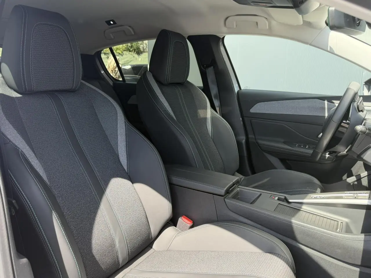 Vue intérieure côté droit de la Peugeot 308 Hybrid 2025 montrant les sièges tissu et cuir gris avec accoudoir central.