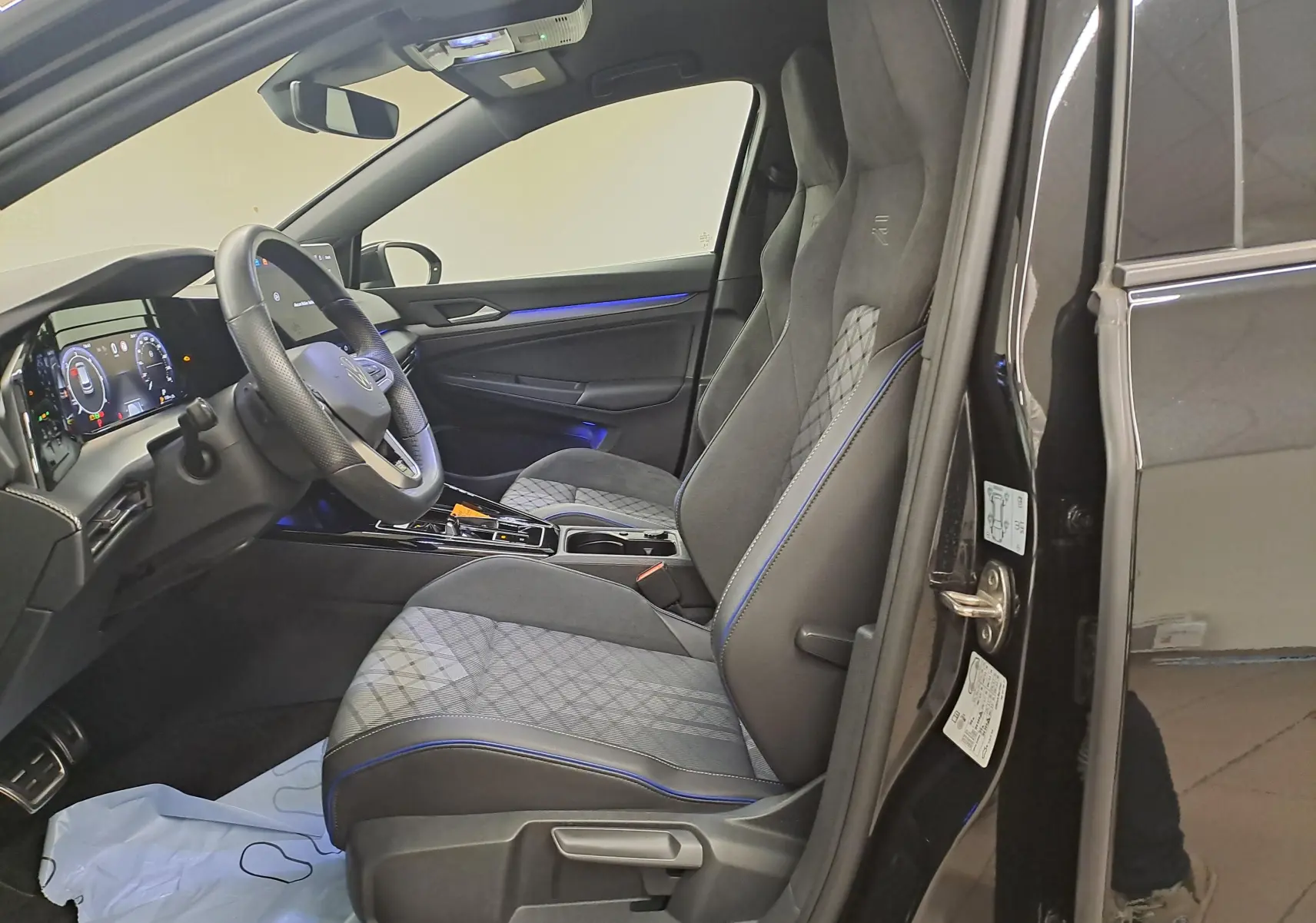 Vue intérieure côté conducteur de la Volkswagen Golf 2.0 TDI R Line 2024 noir grenadille avec sièges sport et éclairage d’ambiance bleu.