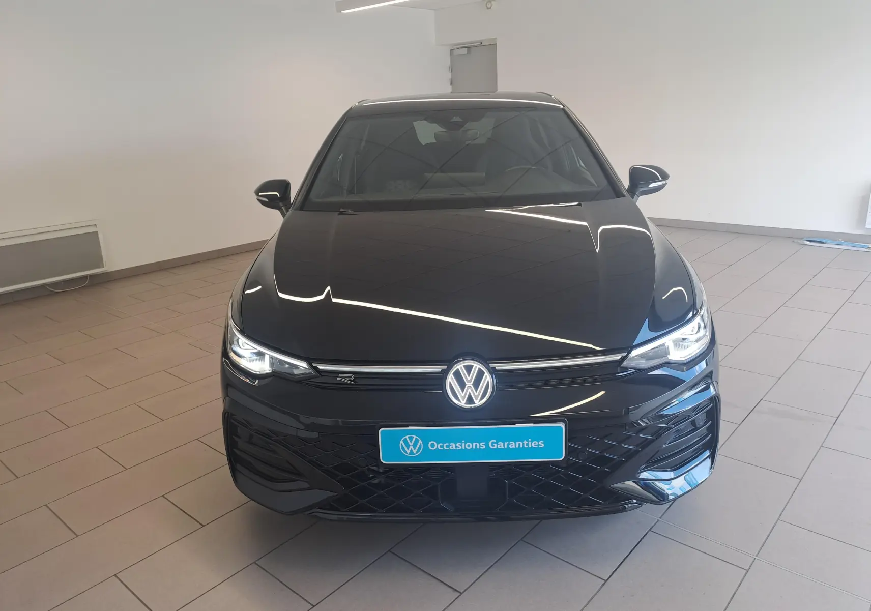 Vue avant d'une Volkswagen Golf 2.0 TDI 150 DSG7 R Line noire grenadille avec phares allumés en intérieur showroom.