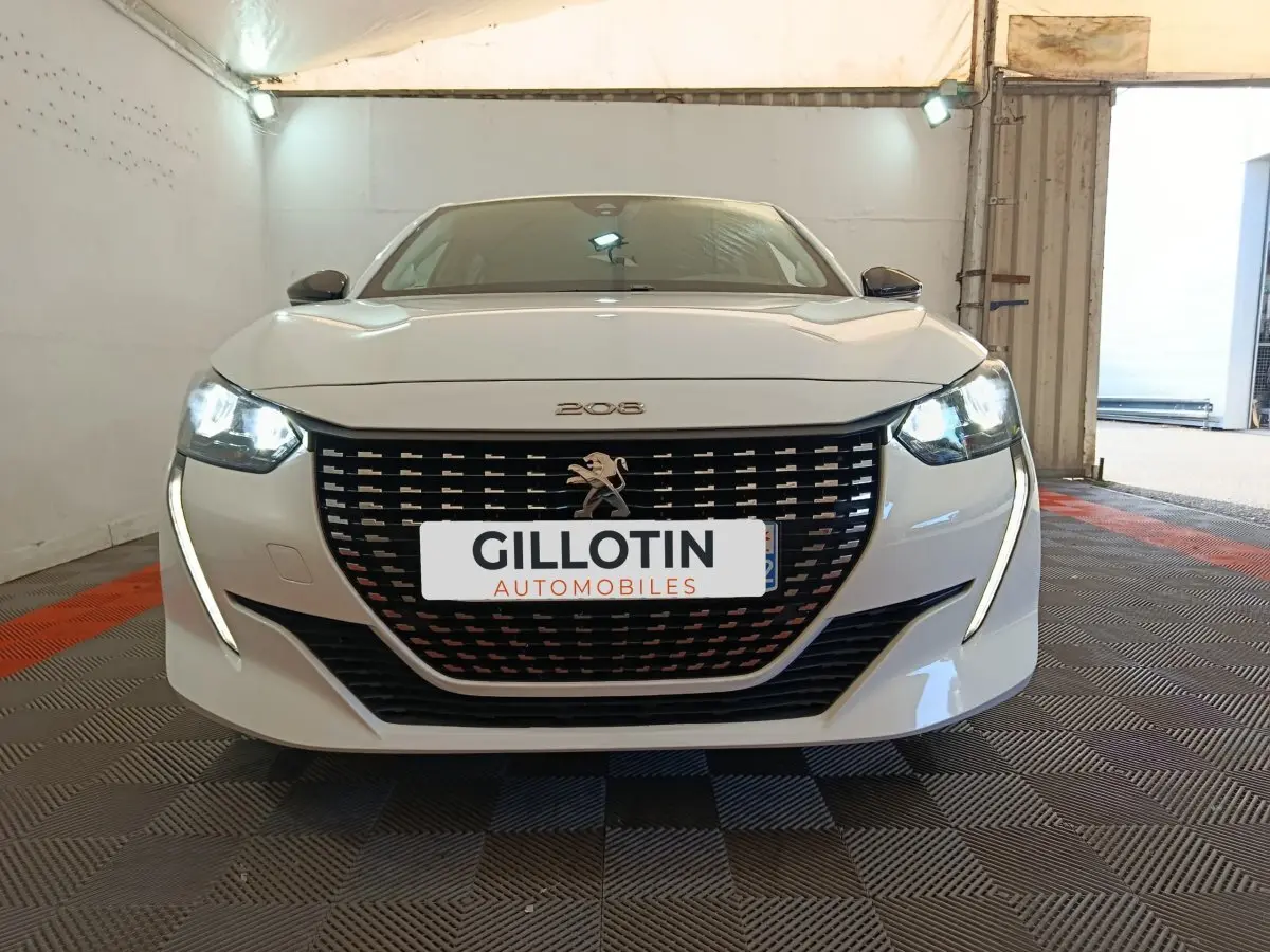 Vue frontale d'une Peugeot 208 blanche 2023 avec phares LED allumés et calandre noire distinctive.
