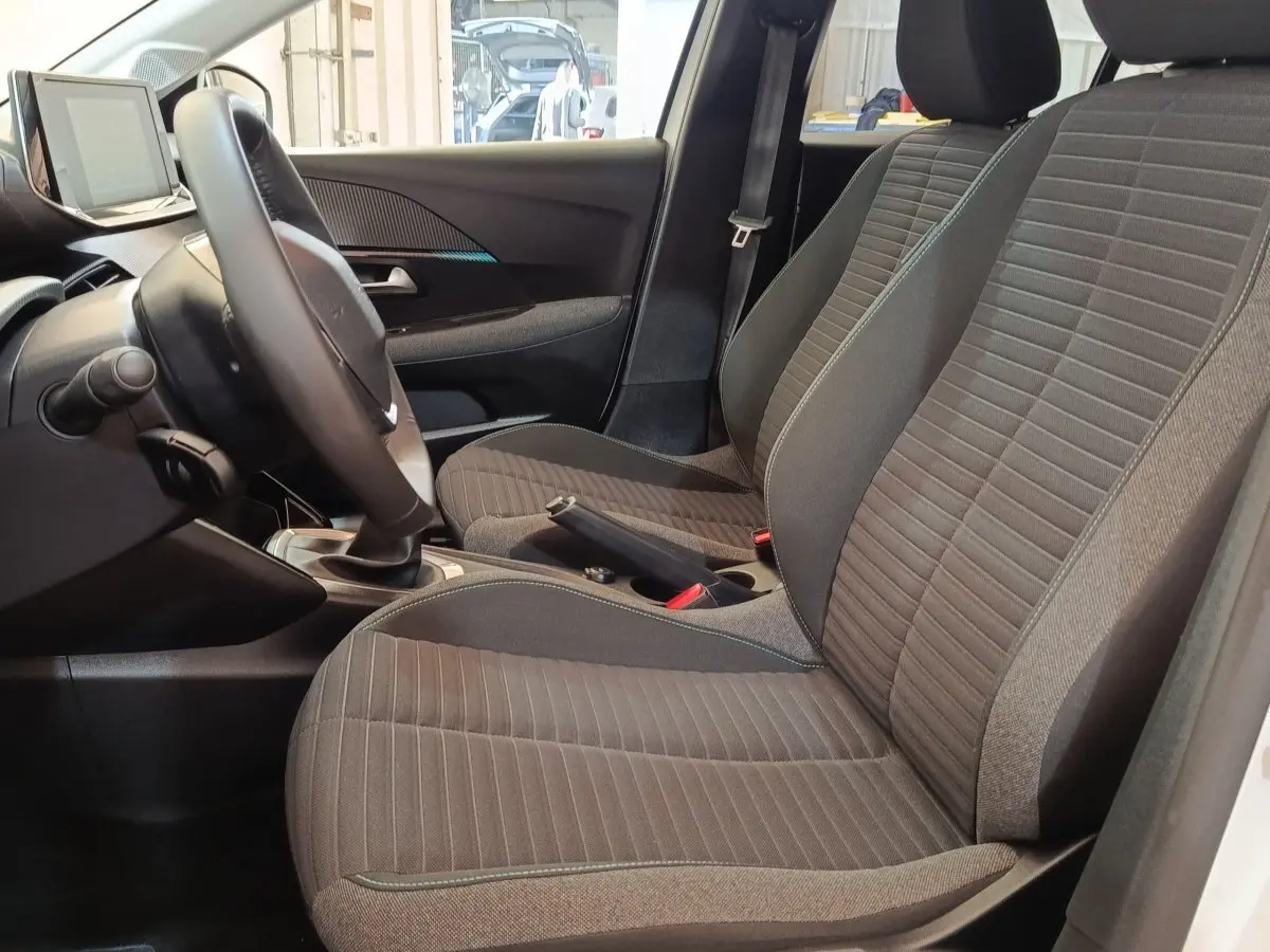 Vue intérieure côté conducteur de la Peugeot 208 blanche 2023, sièges tissu gris et volant cuir multifonctions.