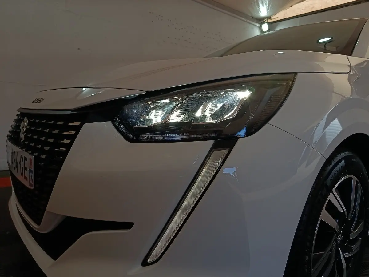 Gros plan sur l'avant droit d'une Peugeot 208 blanche 2023, mettant en valeur son phare LED et la calandre distinctive.