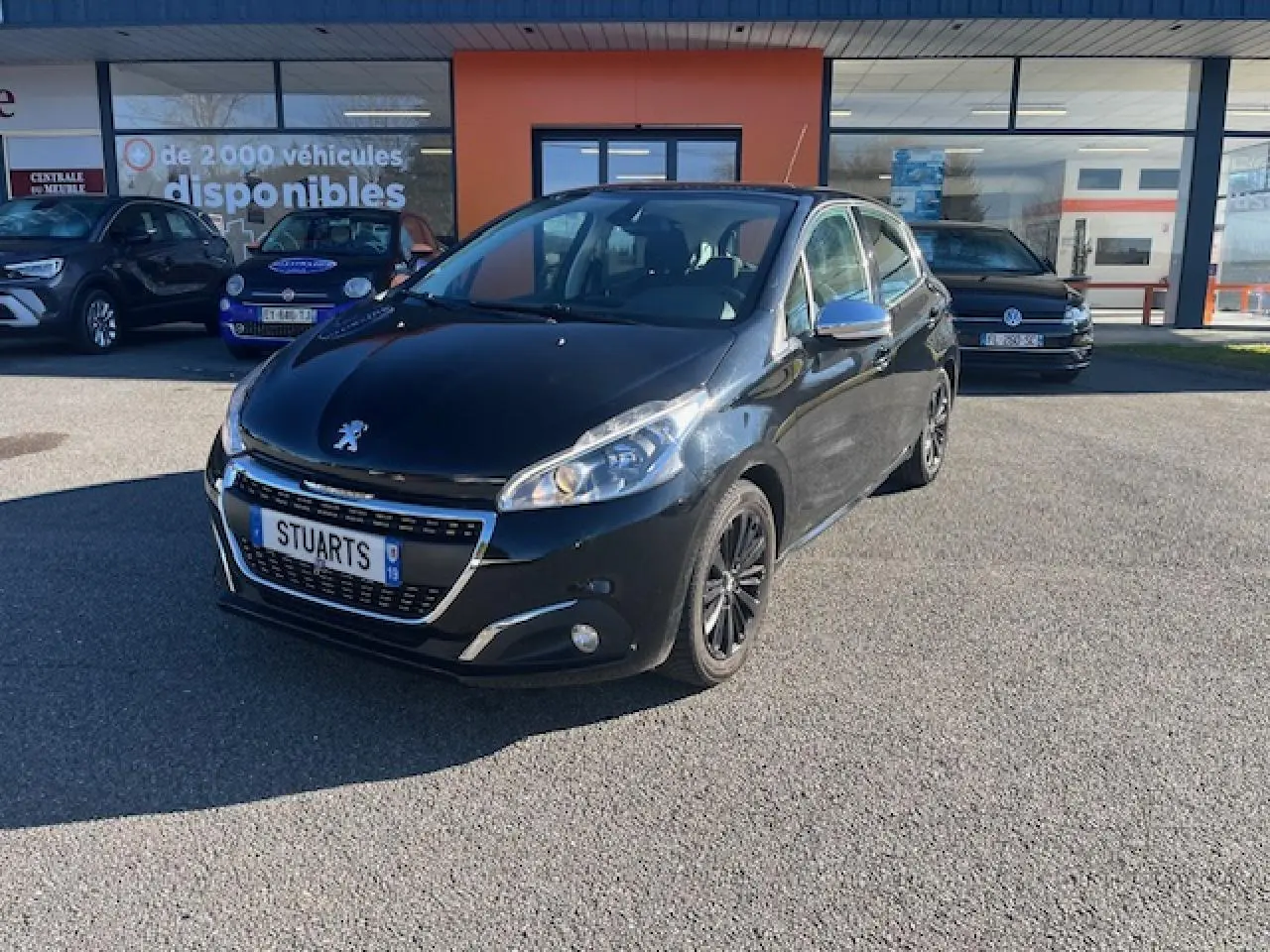 Peugeot 208 noire vue 3/4 avant droit, avec jantes noires et calandre chromée, stationnée devant un bâtiment commercial.